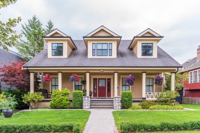 A tan house with red door showcases hanging flowers on the porch, surrounded by greenery and neighboring homes.