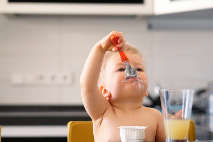 Adorable one year old baby boy eating yoghurt with spoon. Dirty messy face of toddler child.