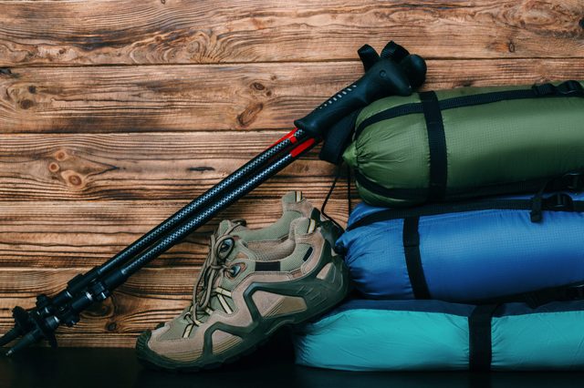 Camping gear rests against a wooden wall; a pair of hiking boots, trekking poles, and stacked sleeping bags in green, blue, and teal.