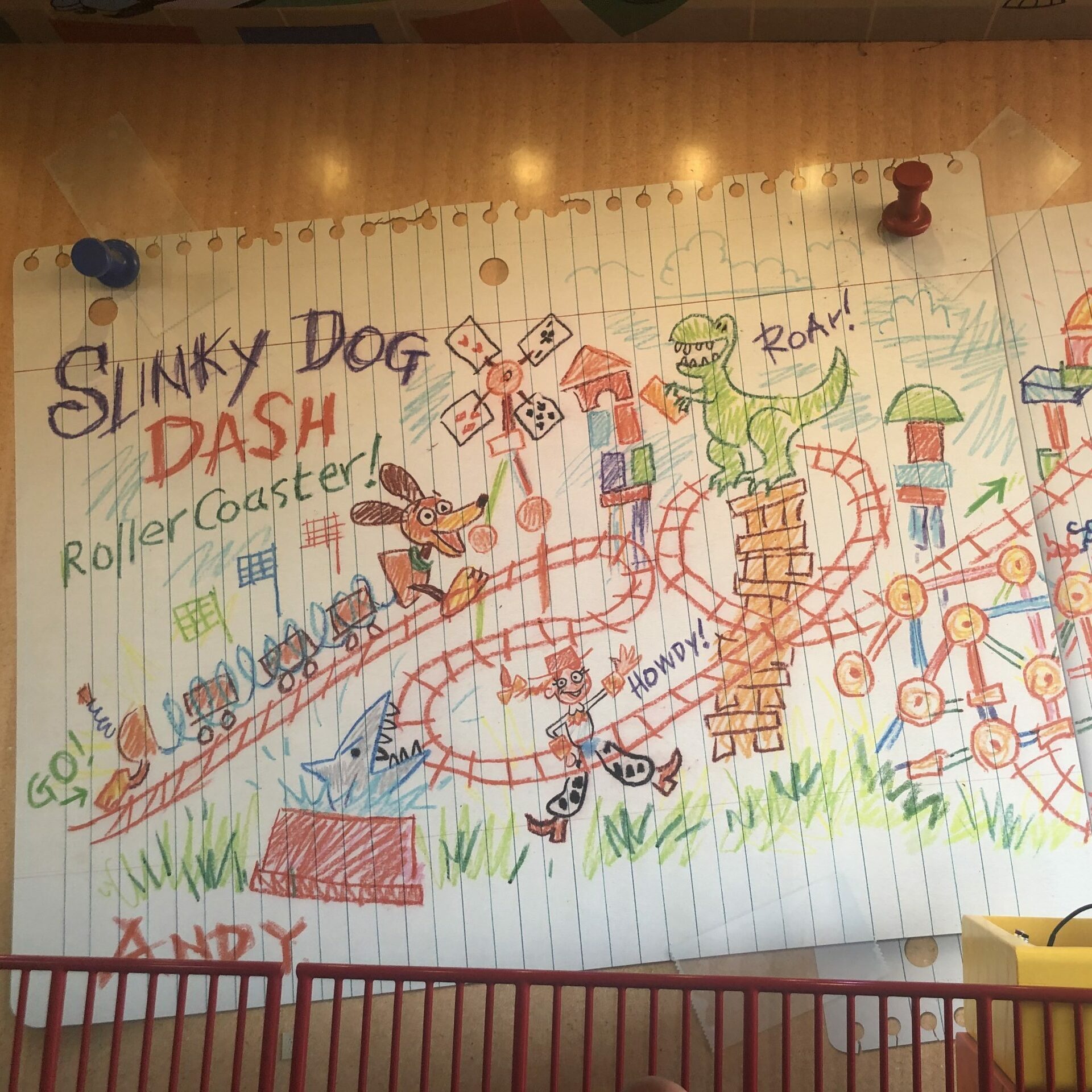Crayon drawing of a roller coaster featuring a dog, dinosaur, and toys on lined paper, with a woman standing below in front of a yellow podium.