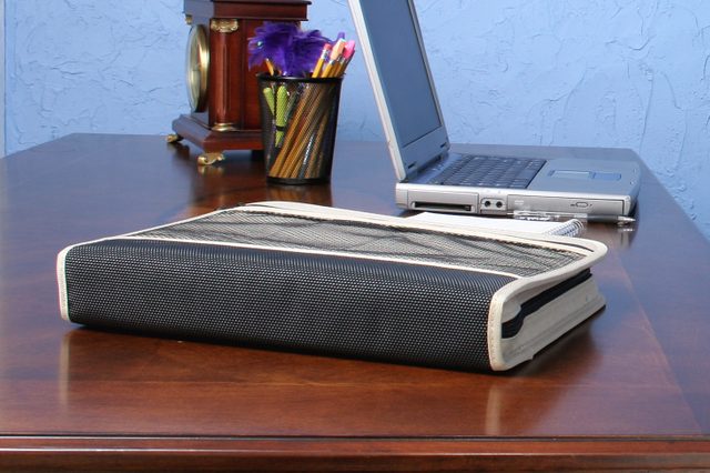 Binder rests on a wooden desk beside a laptop and pencil holder, against a blue textured wall.