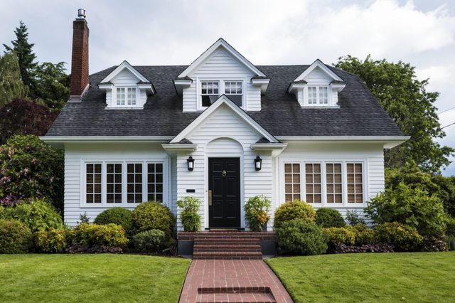 A white house with a black door and chimney sits amidst lush greenery, featuring a brick path and manicured lawn.