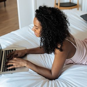Person typing on a laptop while lying on a bed in a cozy bedroom setting.