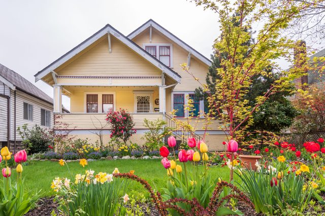 A yellow house stands with colorful flowers blooming in the lush front garden, surrounded by neighboring homes under a cloudy sky.
