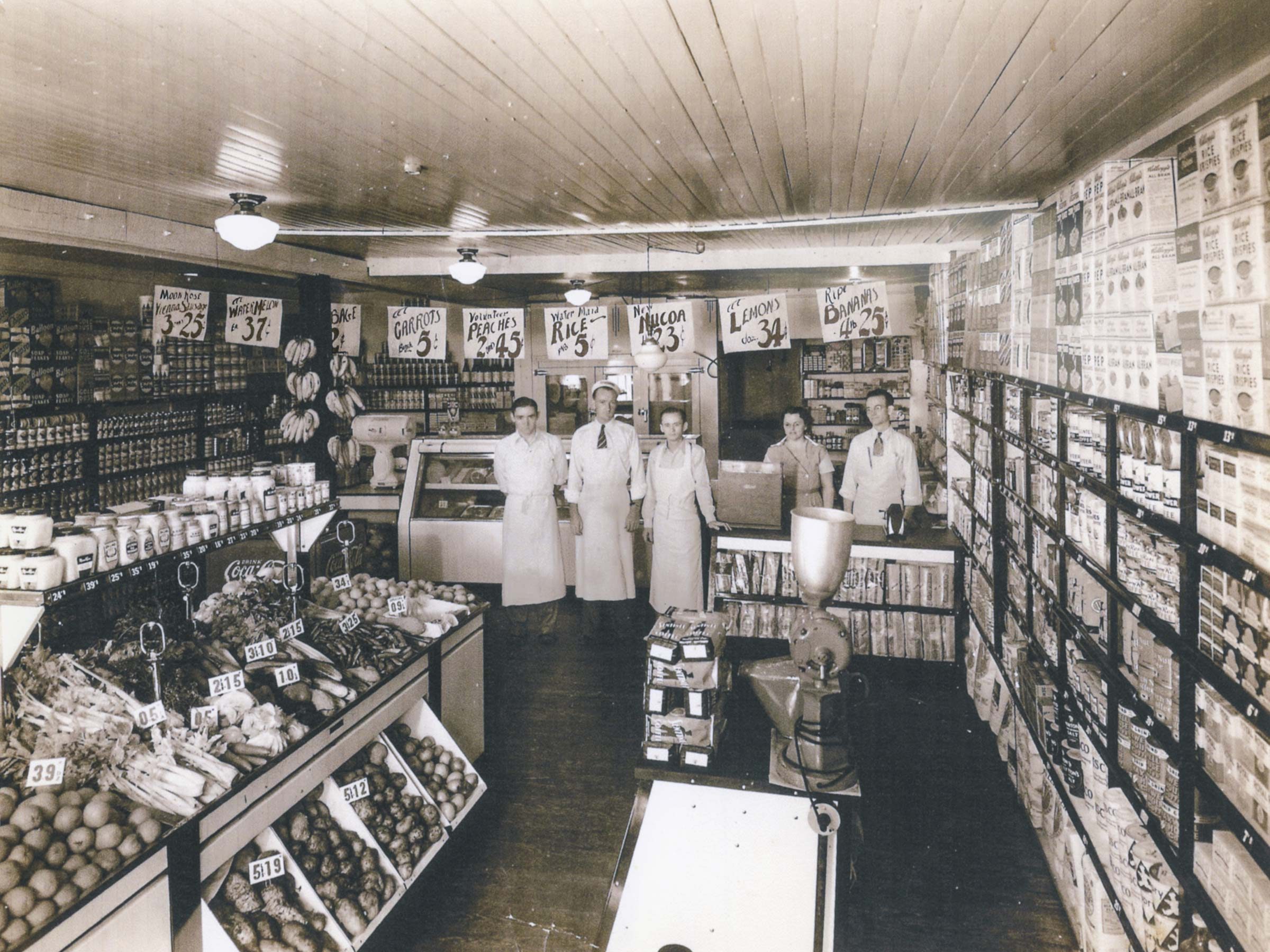 Five people stand in a vintage grocery store, surrounded by shelves of canned goods and produce, with handwritten price signs overhead.