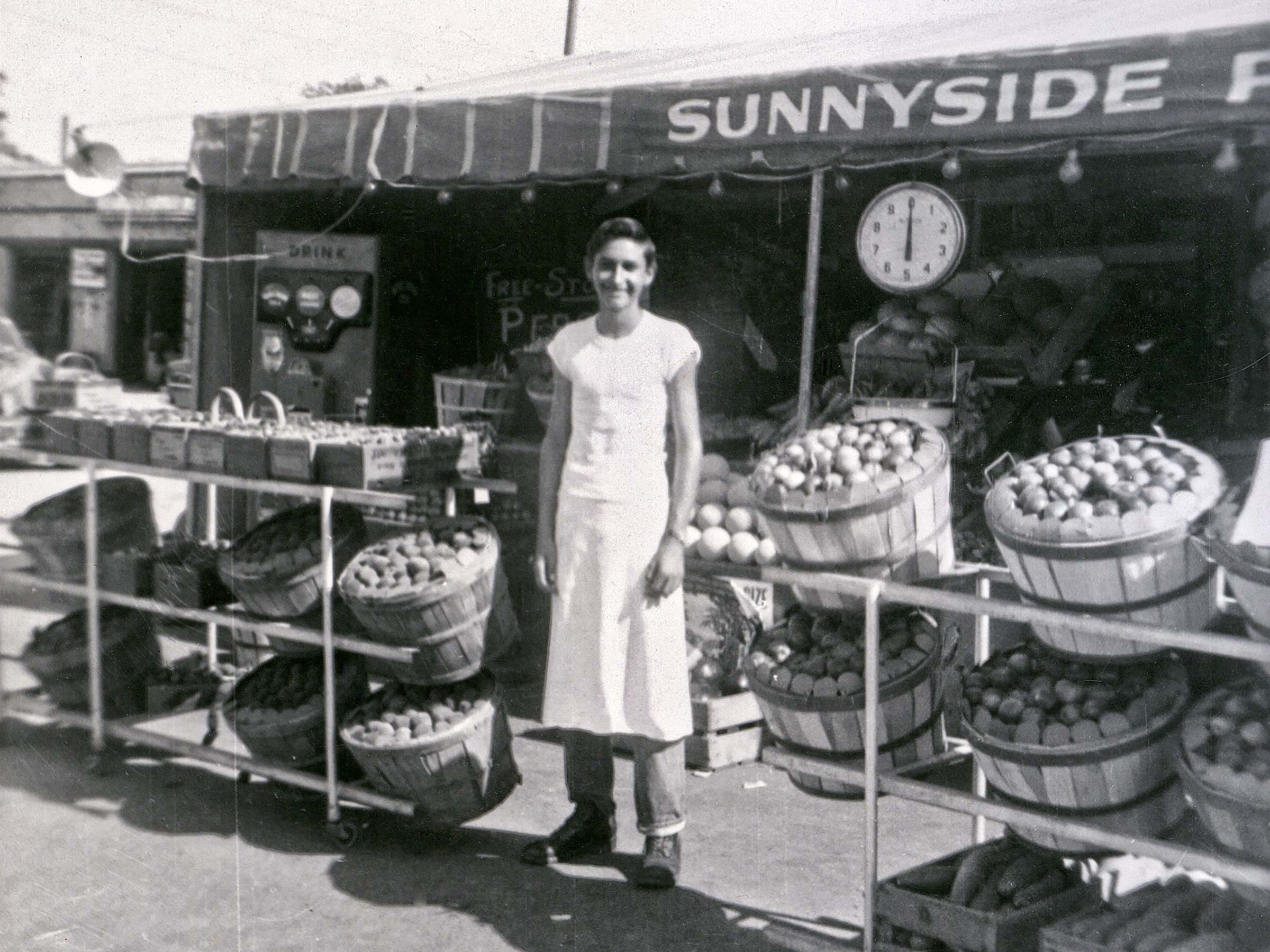 A person stands smiling, wearing an apron, in front of a produce stand with baskets of fruit and a "SUNNYSIDE" awning overhead.