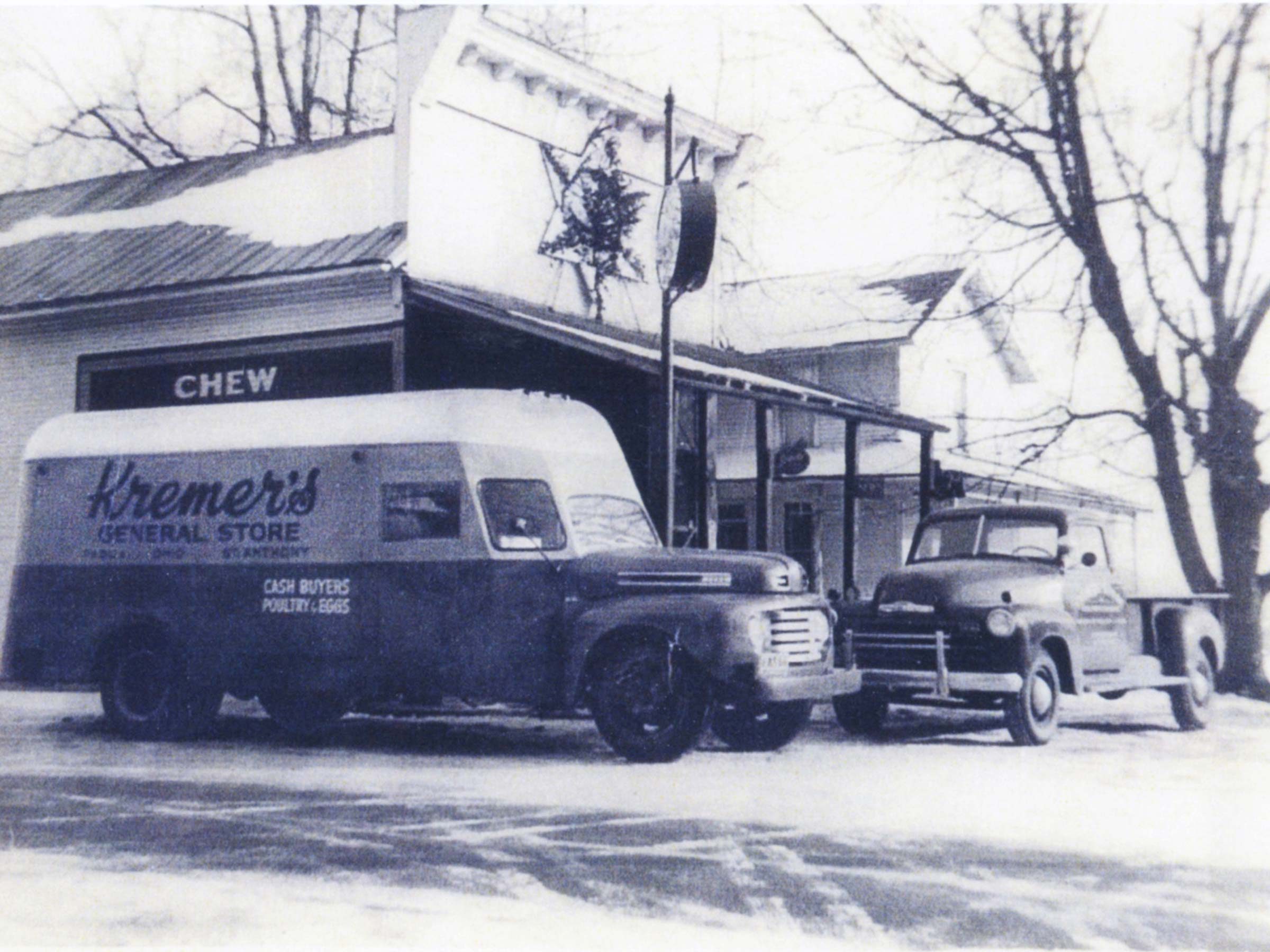 Two vintage trucks parked outside a general store in a snowy rural setting, surrounded by bare trees. Text: "Kremer's General Store" and "CHEW."