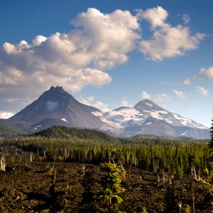Mountains loom under a partly cloudy sky, surrounded by dense forest and rugged lava fields in a natural landscape.