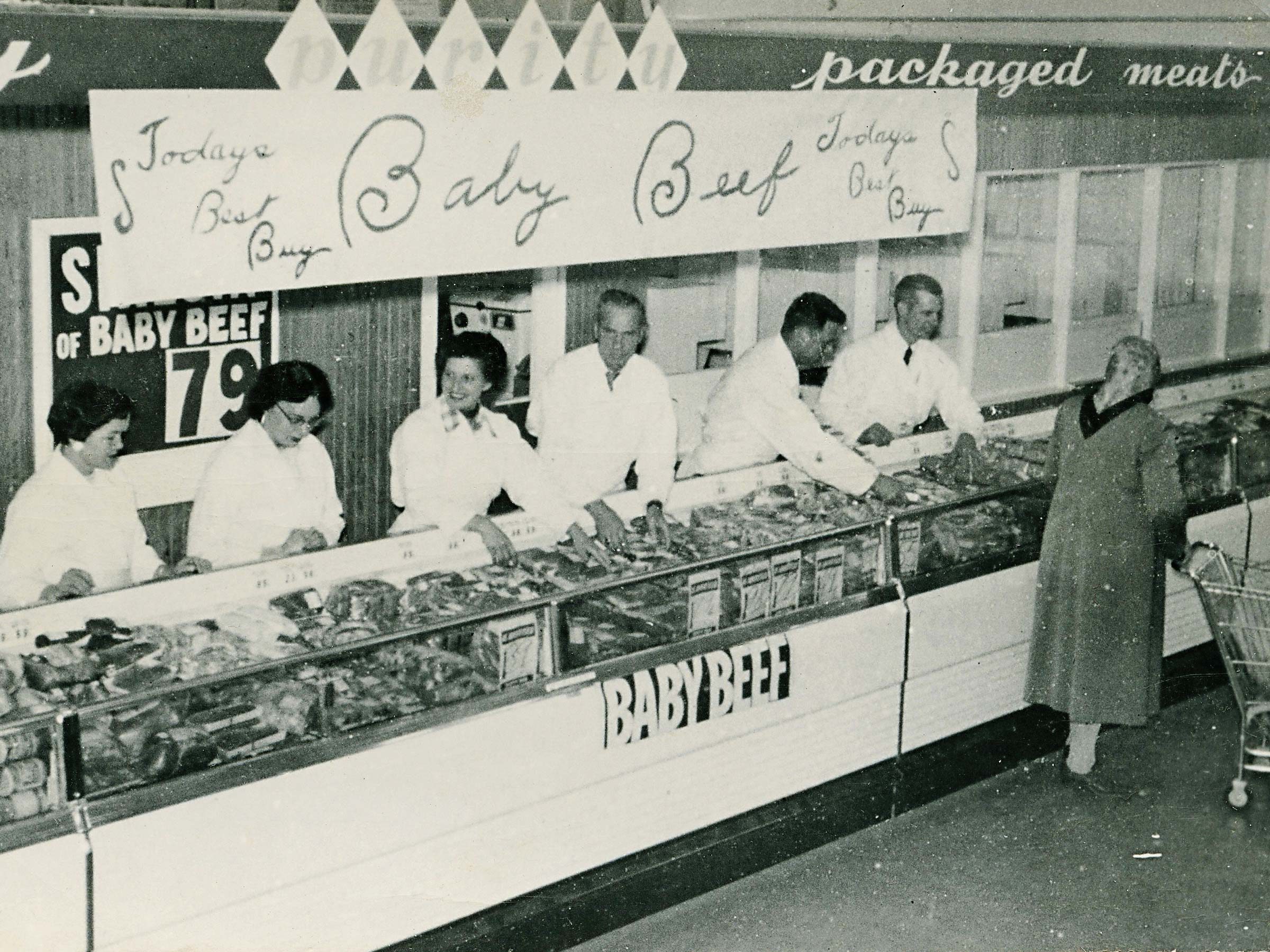 Butchers display meat behind a counter in a supermarket while a customer with a shopping cart looks on. A sign reads "Today's Best Buy Baby Beef."