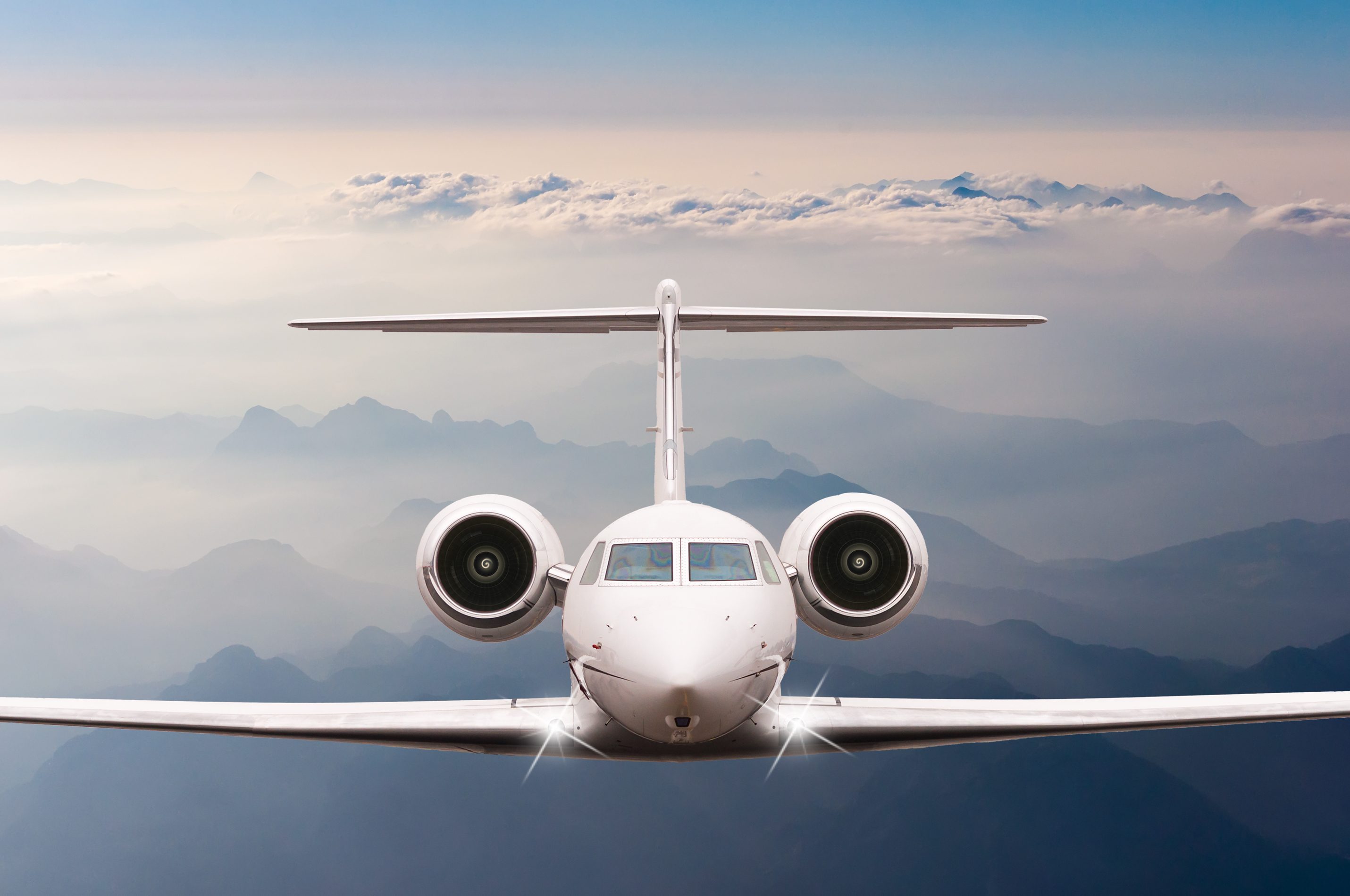 Airplane flying through the sky, viewed head-on, with mountains and clouds visible in the background.