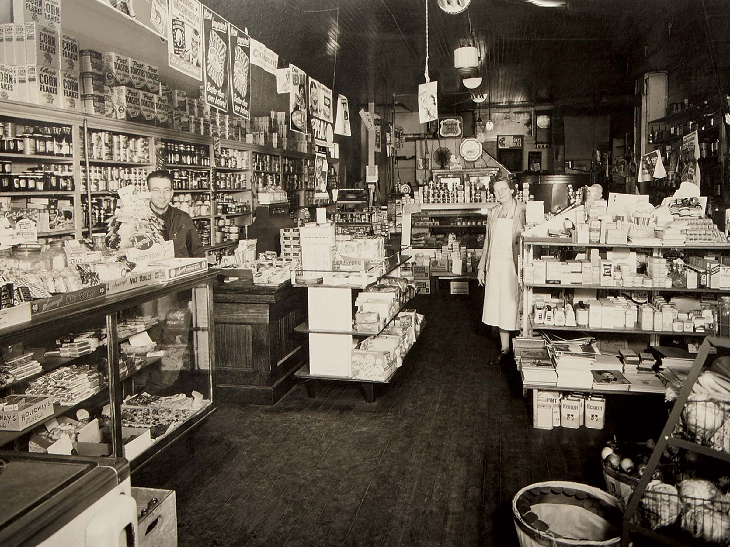 A man and woman stand behind counters, surrounded by stocked shelves, in a vintage grocery store filled with various canned goods and products.