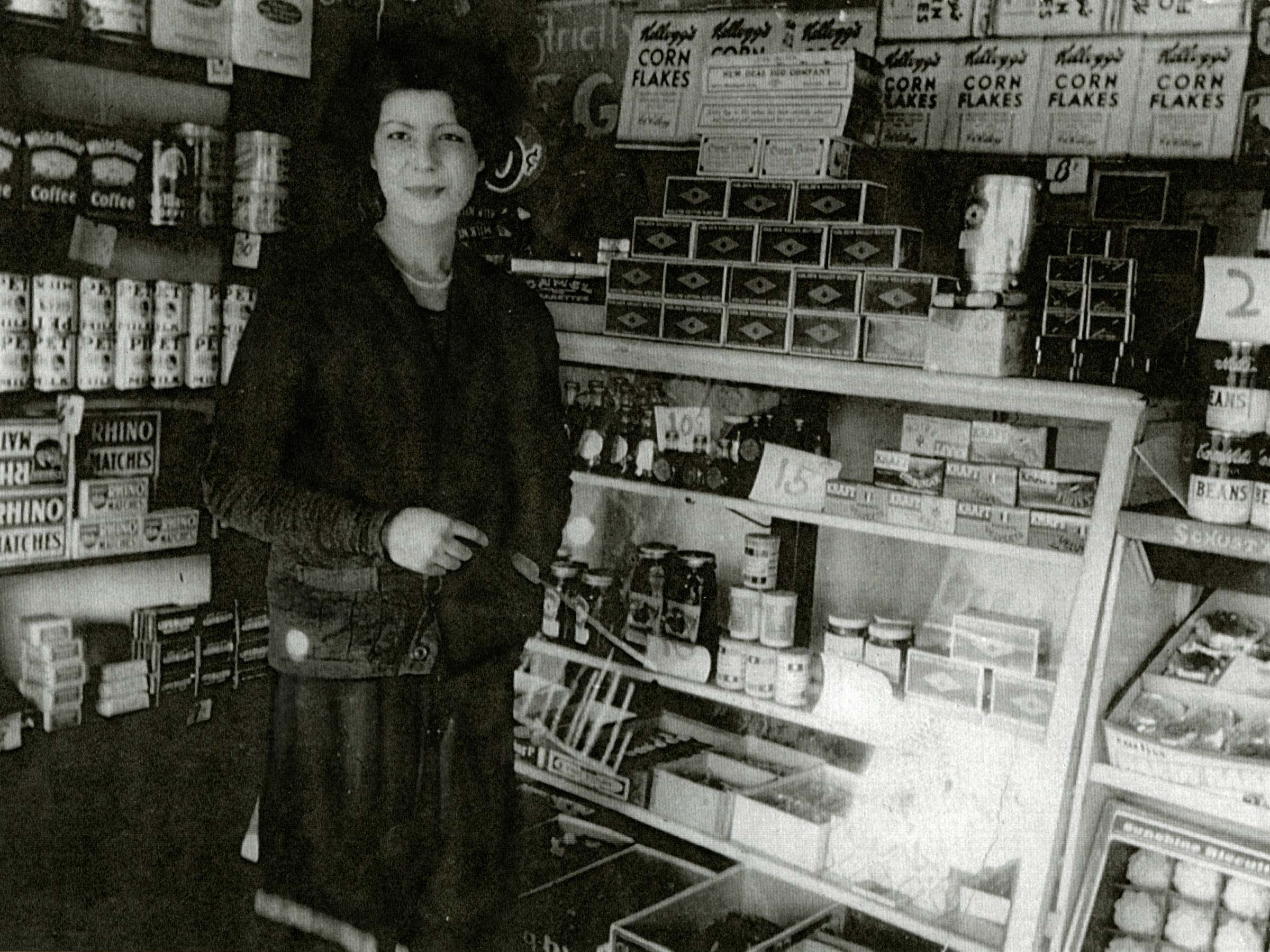 A woman stands in a small shop filled with shelves of canned goods, corn flakes boxes, matchboxes, and other products.