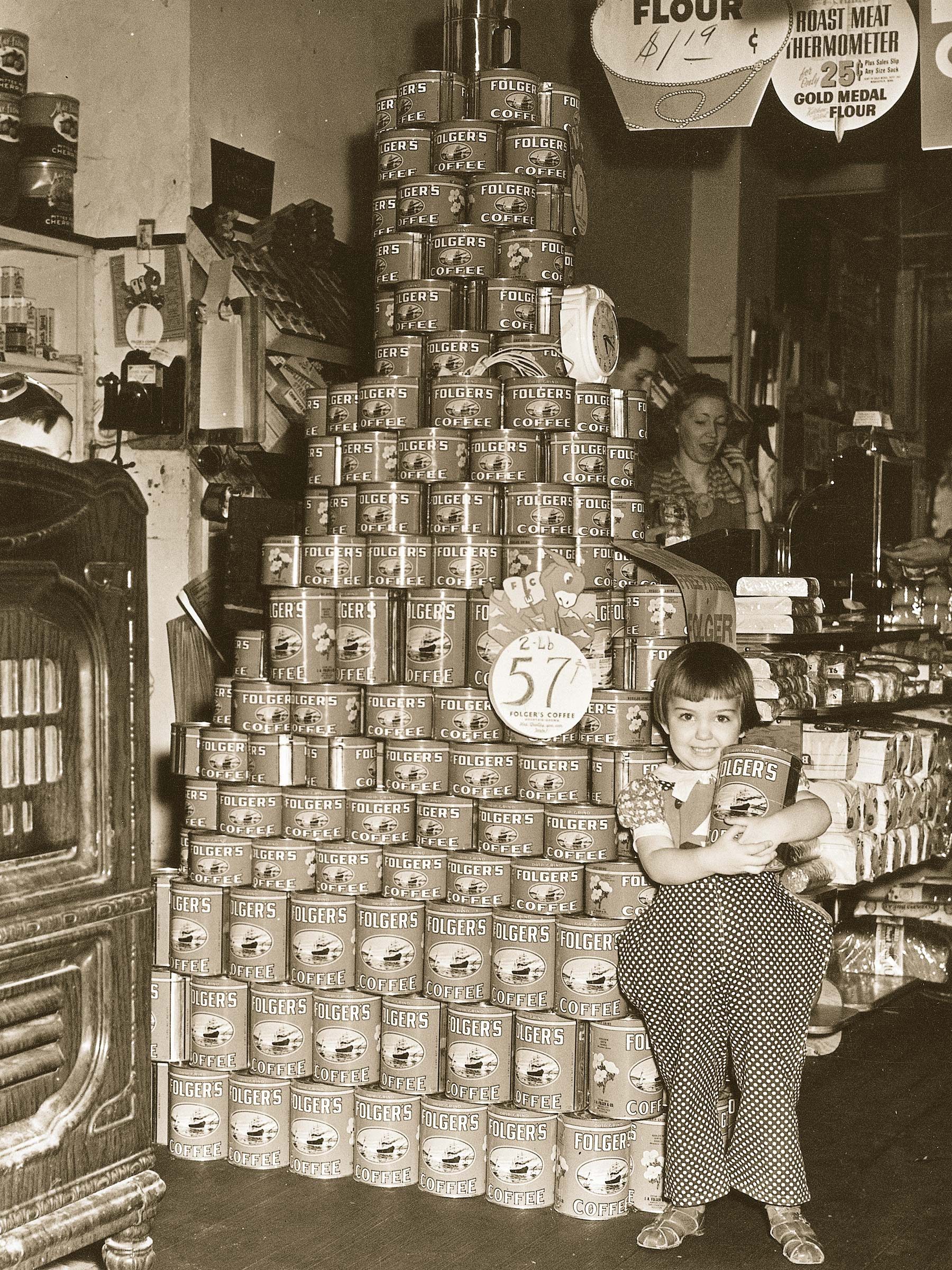 A child hugs a coffee can, standing before a large pyramid of stacked Folgers Coffee cans, inside a vintage store setting.