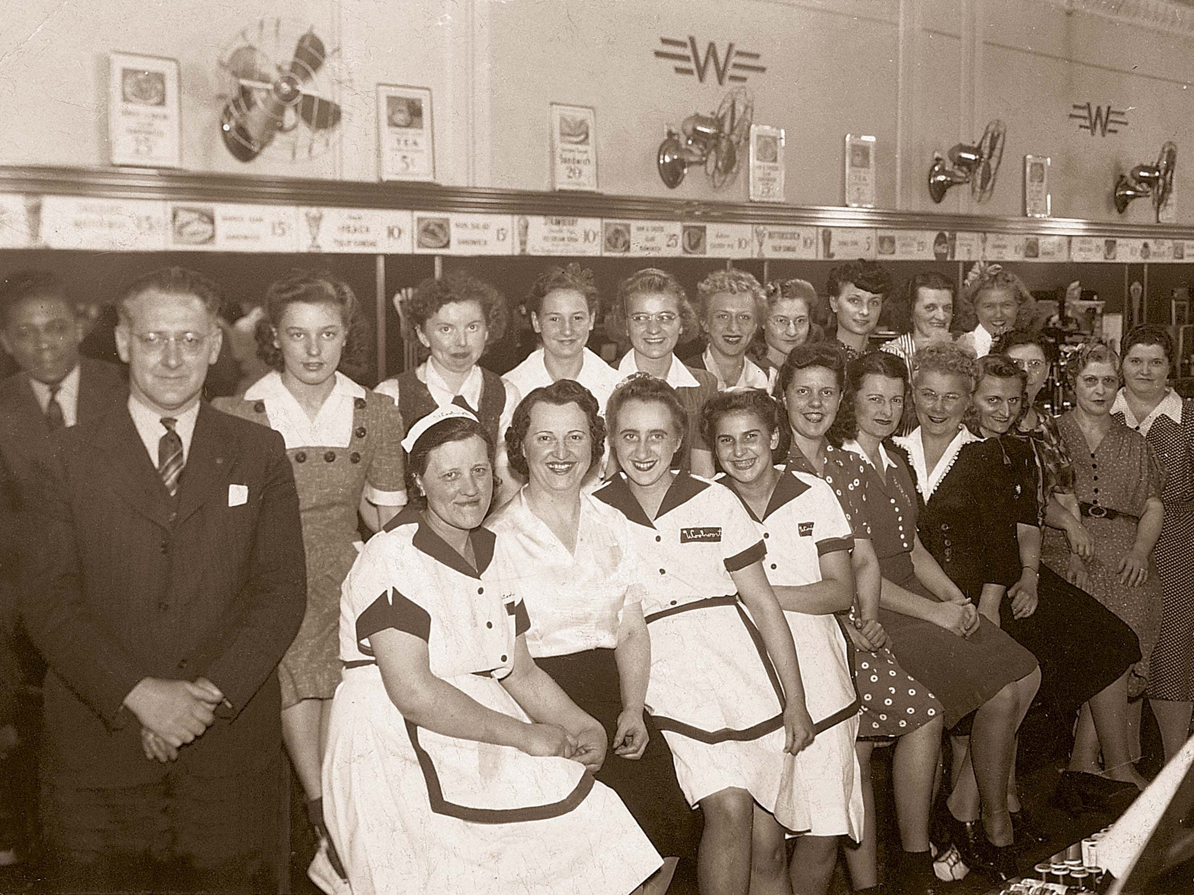 A group of people, mostly women in uniforms, smiling together indoors. The setting appears to be a diner or restaurant with visible wall fans.