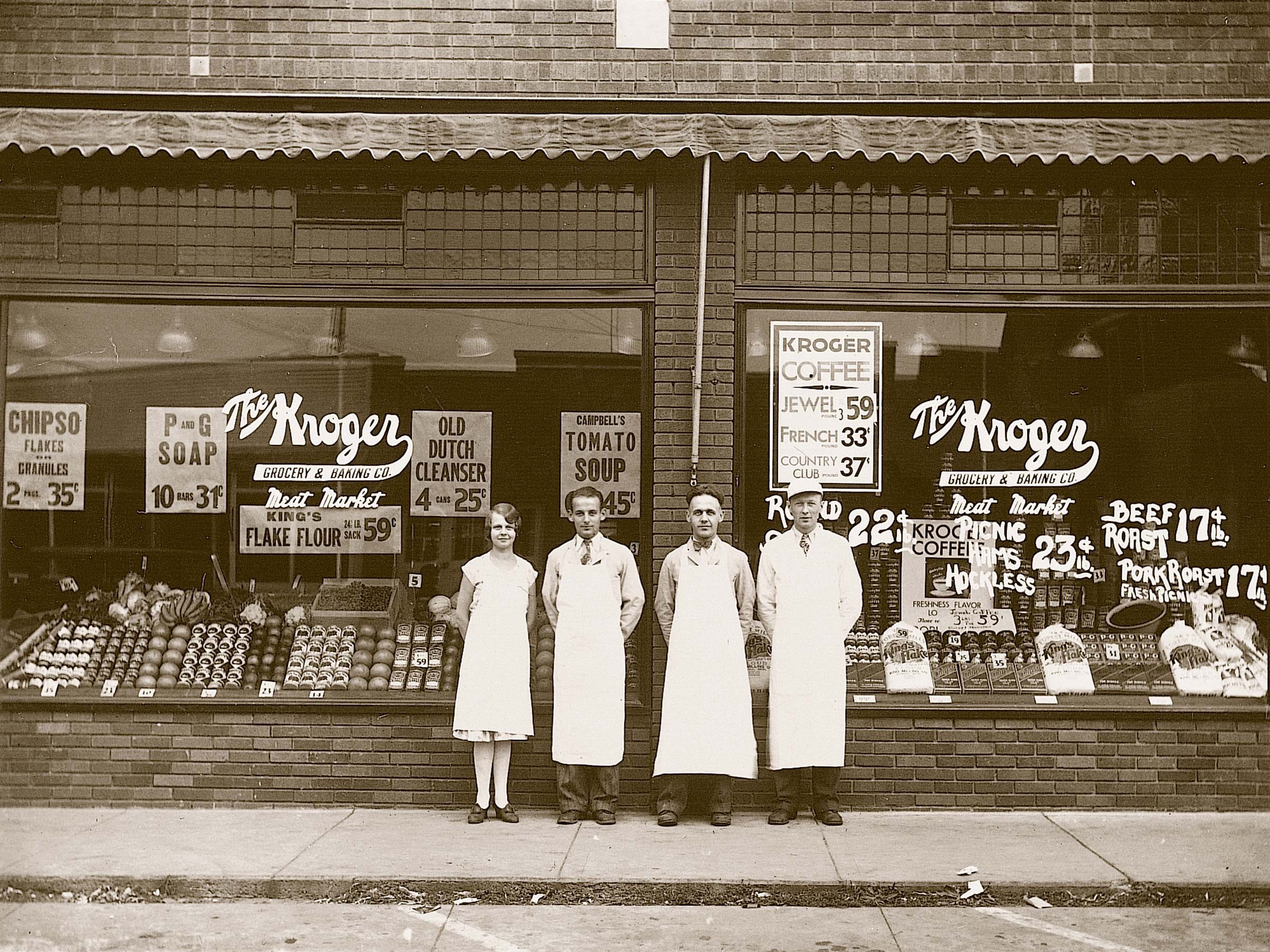 Four people stand in front of a brick storefront with glass windows displaying groceries and prices, under an awning reading "The Kroger Grocery & Baking Co."