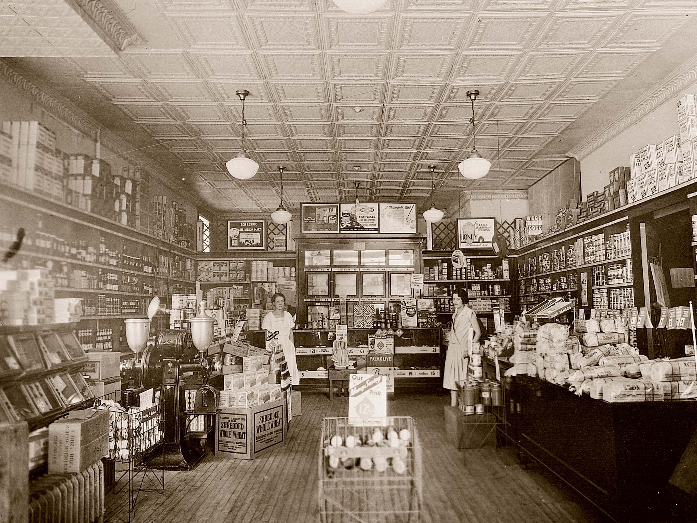 Store interior displays products on shelves, counters; two people assist and organize, surrounded by vintage advertisements and neatly arranged grocery items.