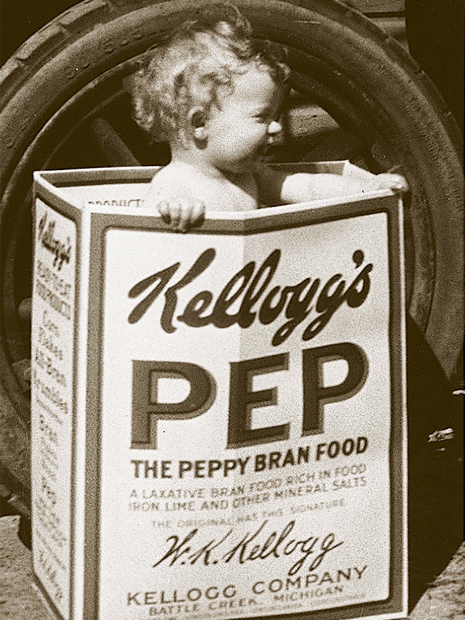 Baby sitting inside a large Kellogg's Pep cereal box, outdoors, beside a large tire wheel.