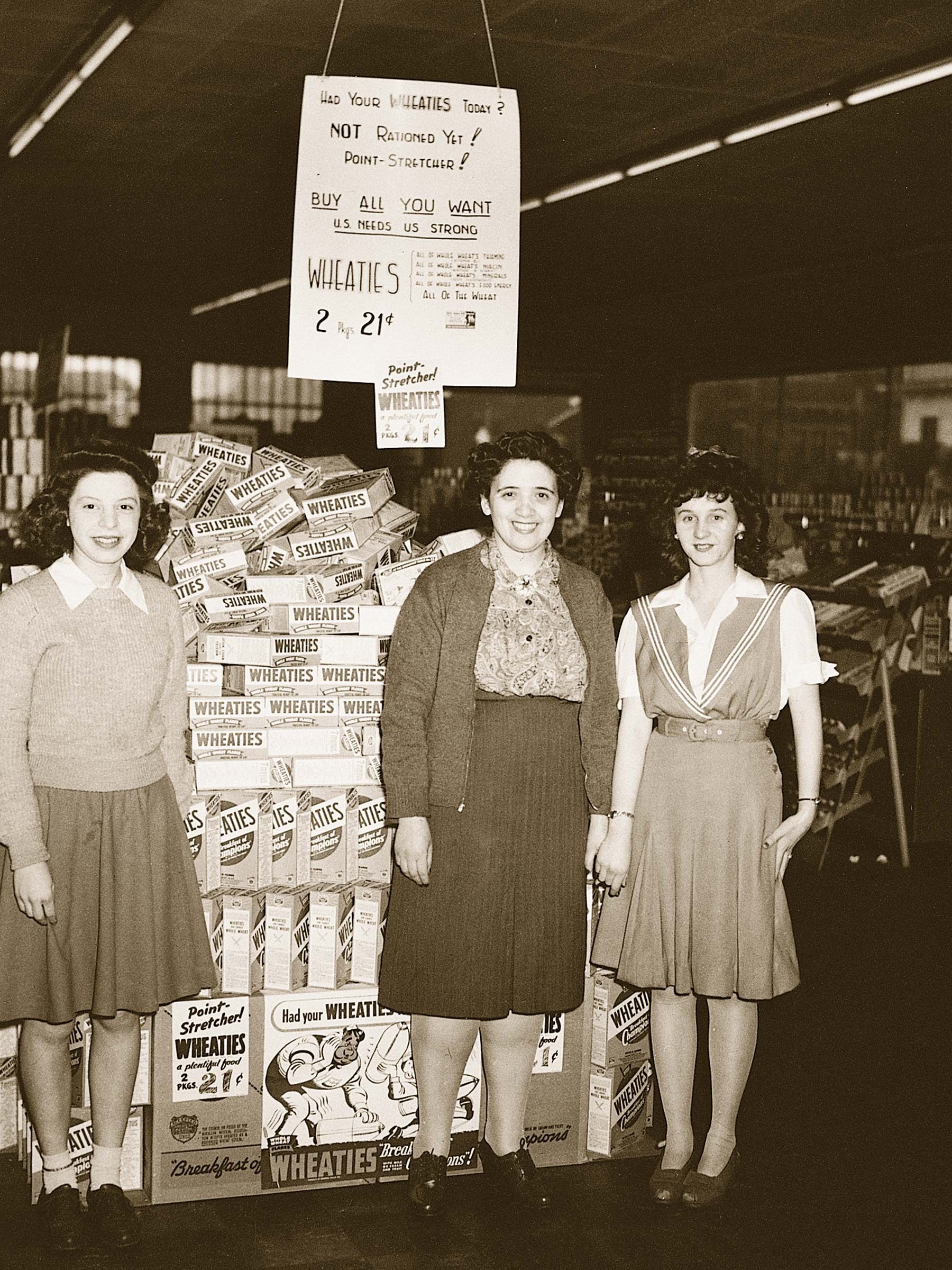 Three women stand smiling in front of stacked Wheaties cereal boxes, inside a dimly lit store, with a promotional sign overhead.