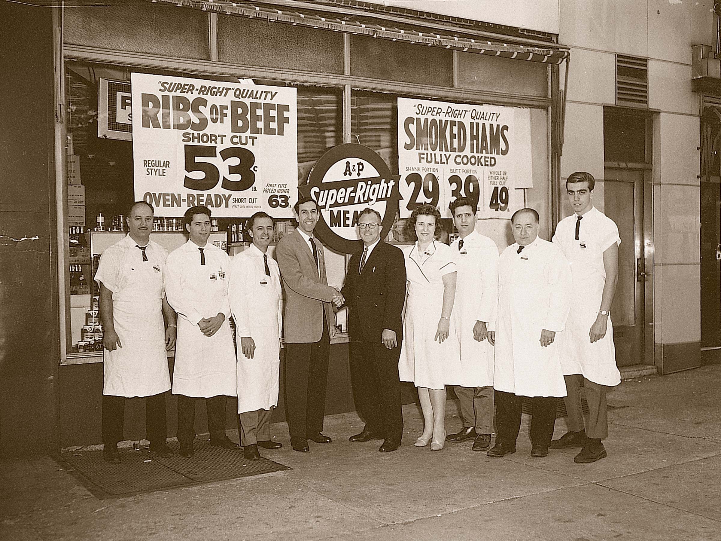 A group of people stands outside a store, wearing uniforms. Two men shake hands. Signs advertise "RIBS OF BEEF" and "SMOKED HAMS."