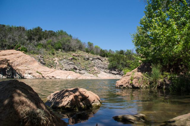 Rocks sit by a calm river, surrounded by lush greenery and hills under a clear blue sky.