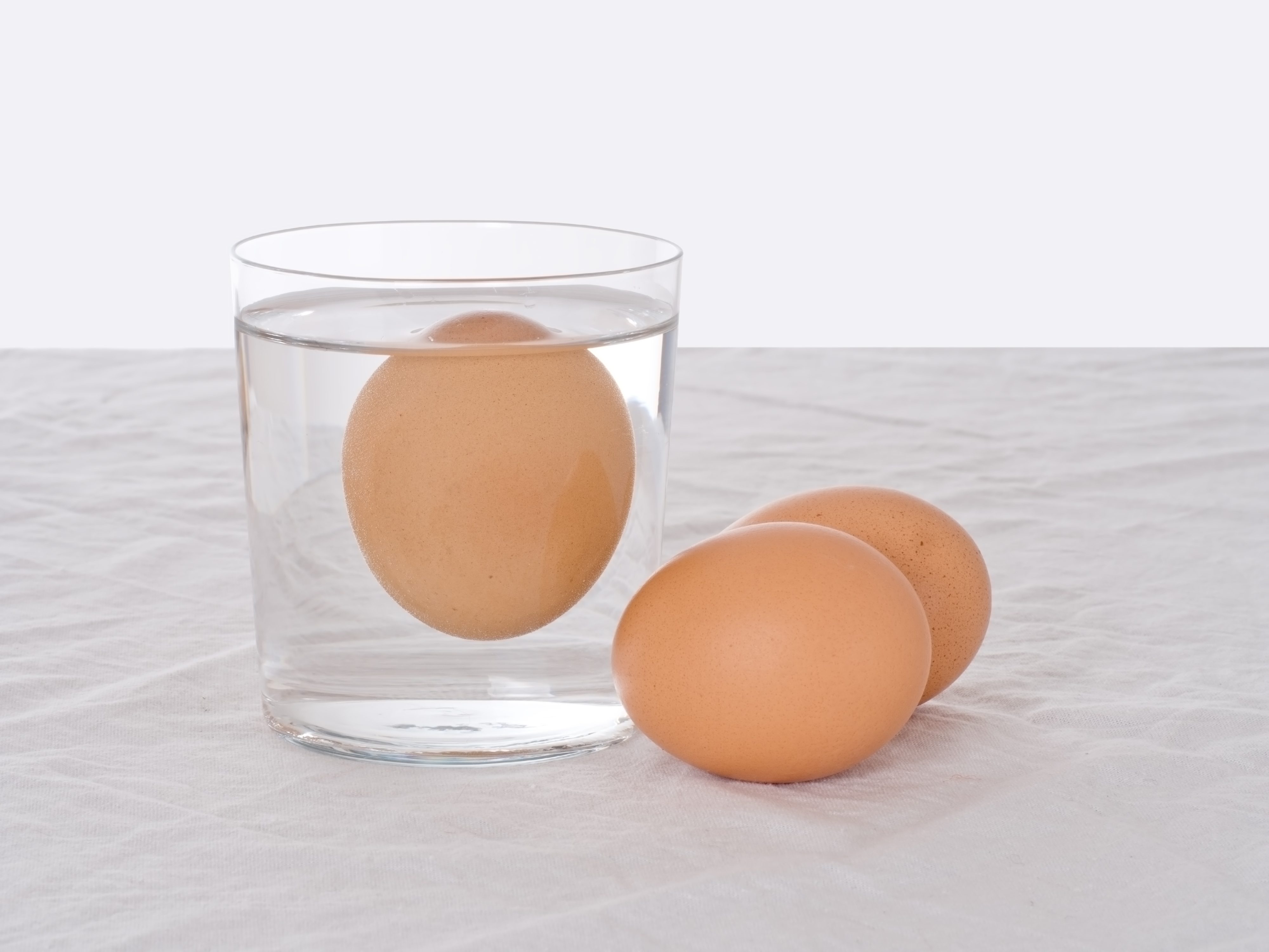 An egg floats in a glass of water; two eggs rest nearby on a white cloth.