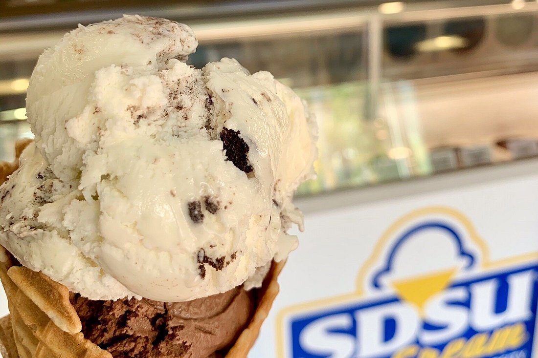 Ice cream cone holds scoops, with vanilla on top and chocolate below, in front of a blurred background displaying â€œSDSU Creameryâ€ signage.