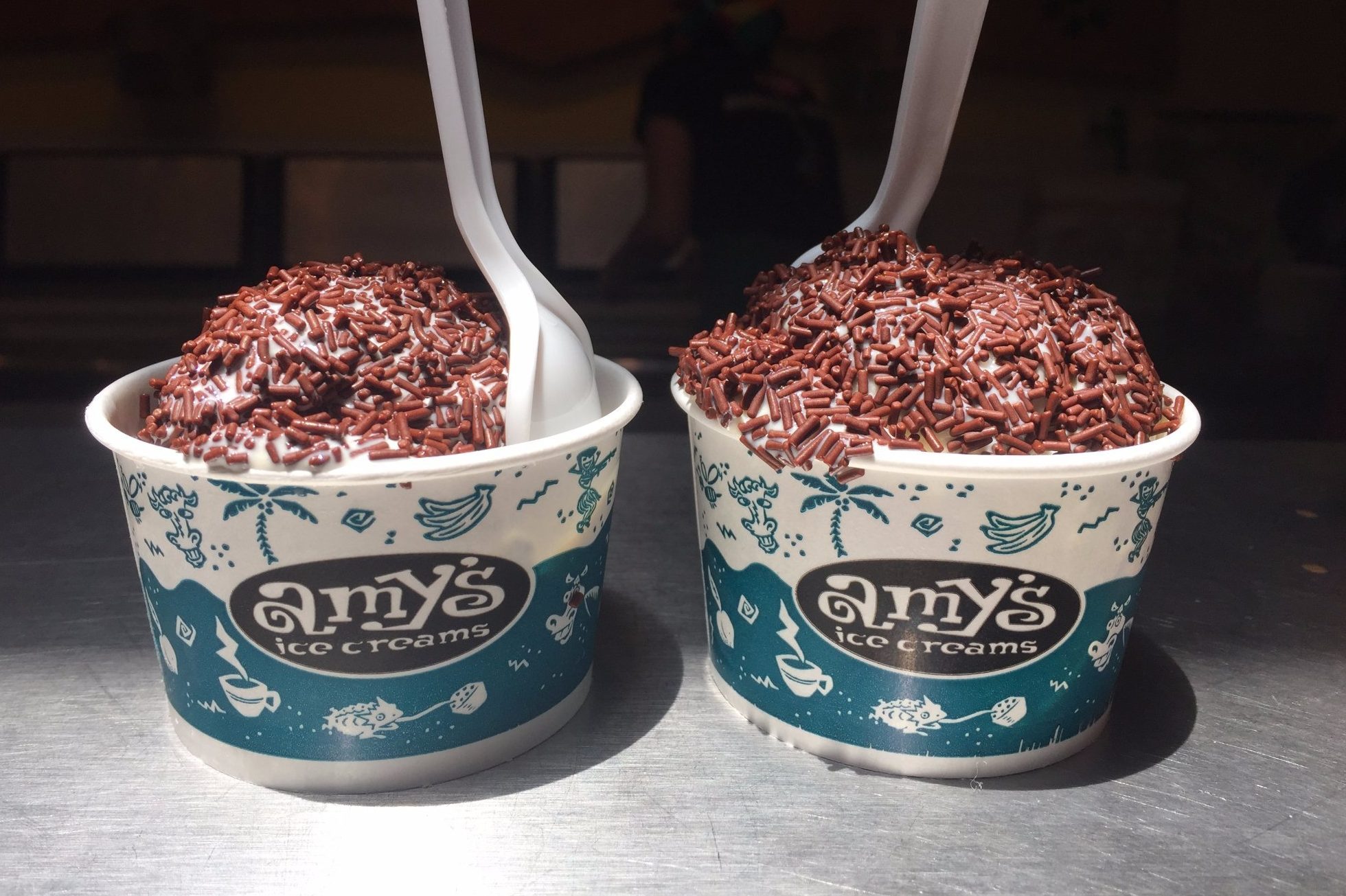 Two cups of ice cream, topped with chocolate sprinkles and spoons, sit on a metal surface. Cups are labeled "amy's ice creams."