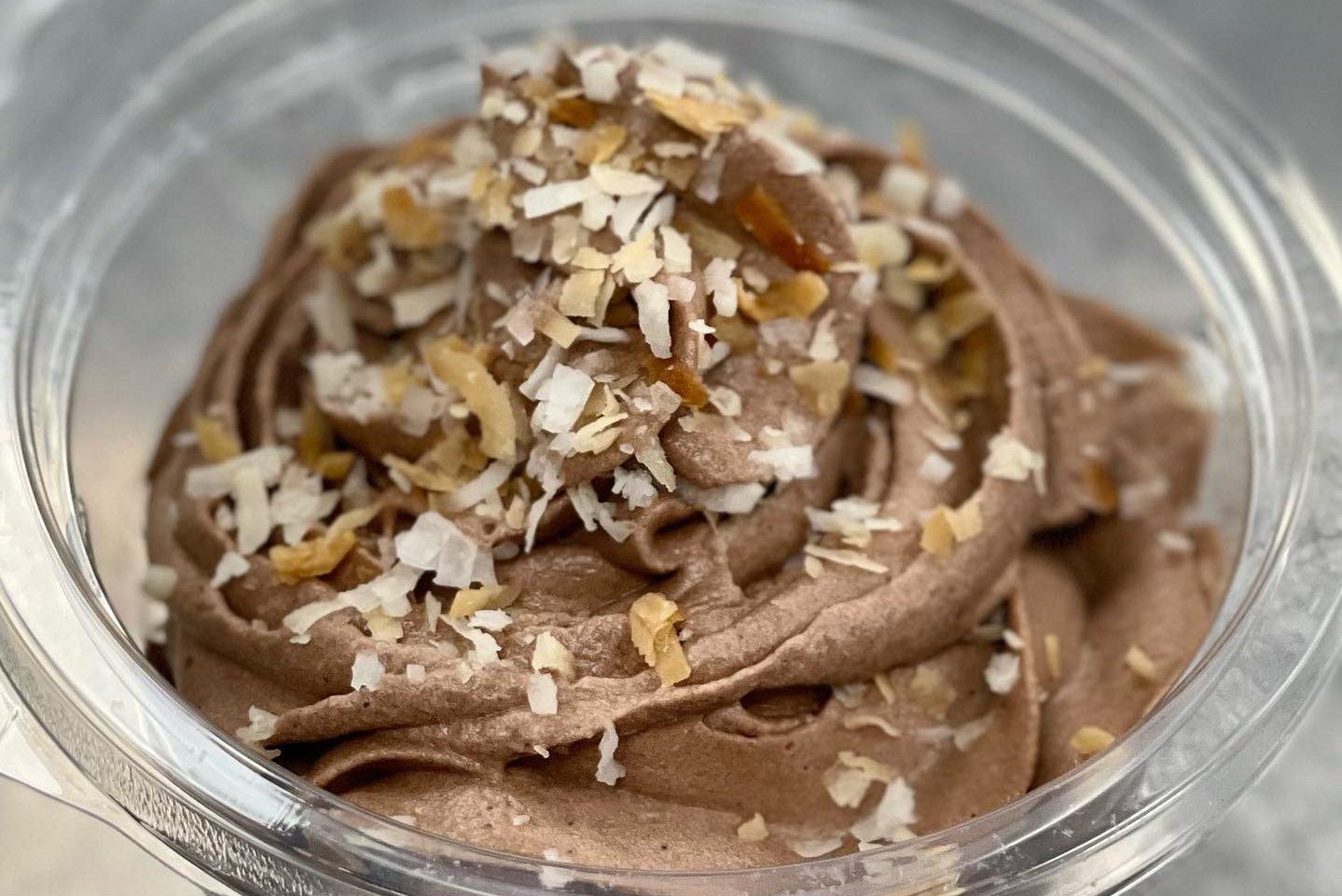 Swirled chocolate mousse topped with shredded coconut and nuts, contained in a clear plastic cup, sitting on a light surface.