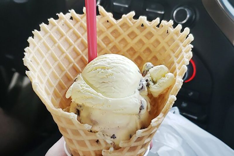 Ice cream rests in a waffle cone, held in hand, with a pink spoon. It is set inside a car with a dark interior.