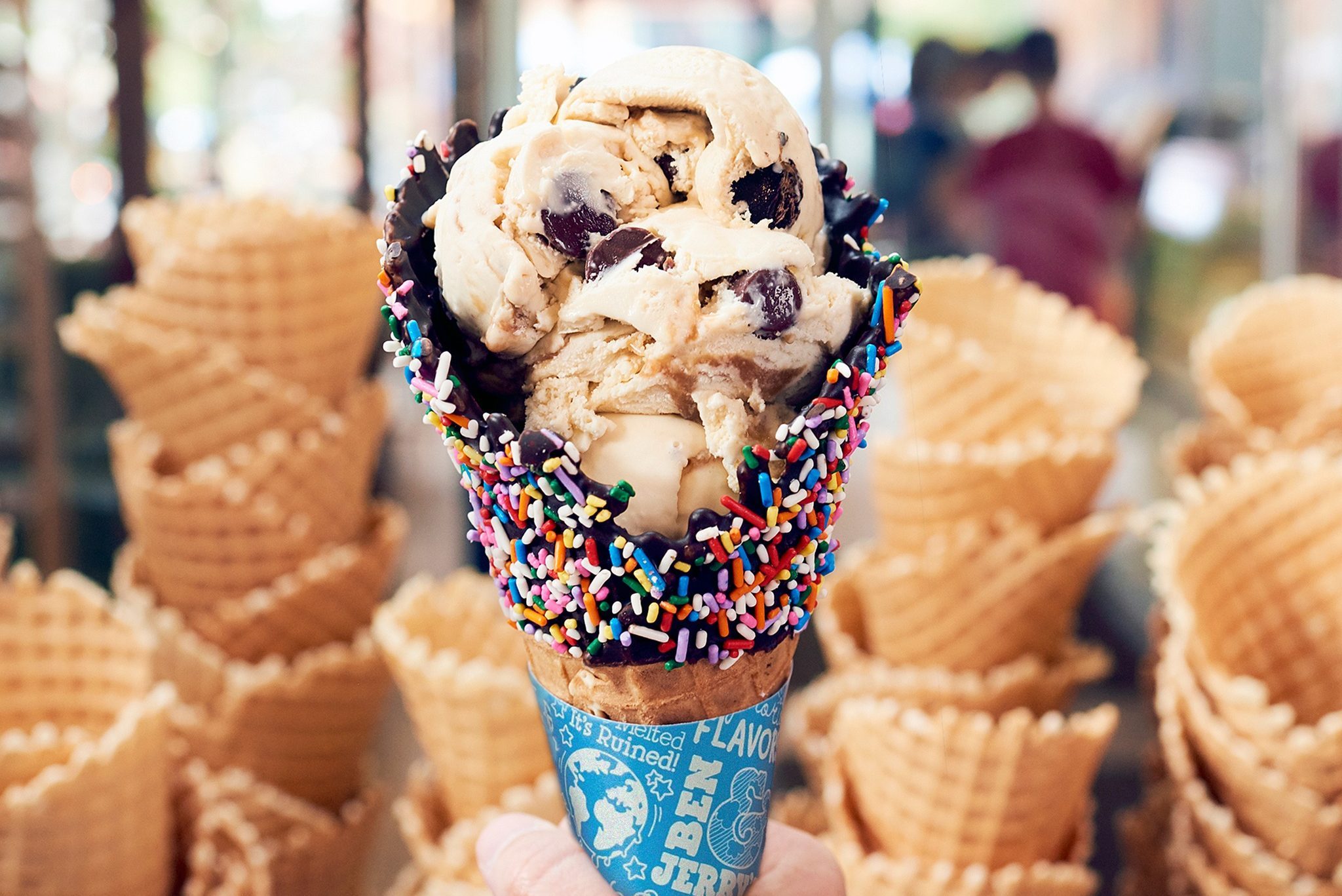 Ice cream scoops rest in chocolate-sprinkle waffle cone, held by a hand; background features stacks of waffle cones in an ice cream shop.