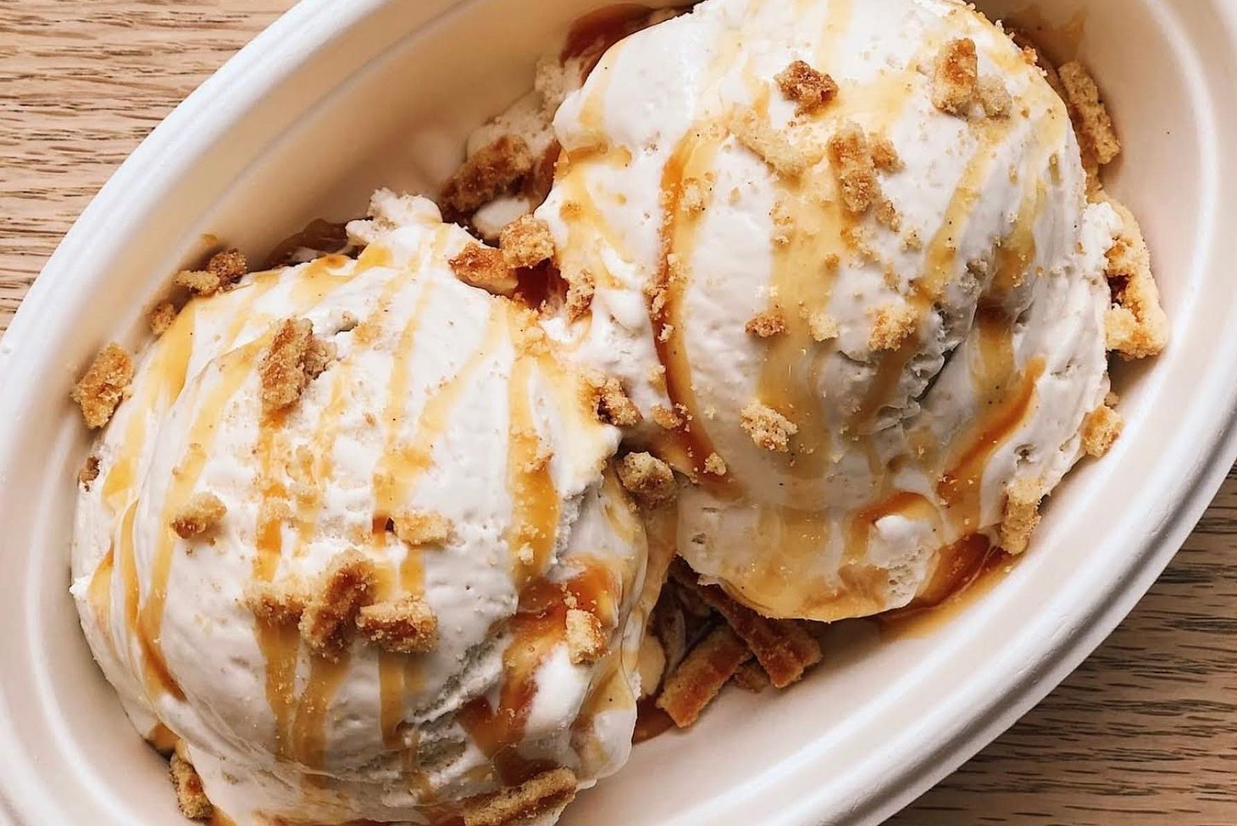 Ice cream with caramel drizzle and crumbled cookies sits in a white bowl on a wooden table.