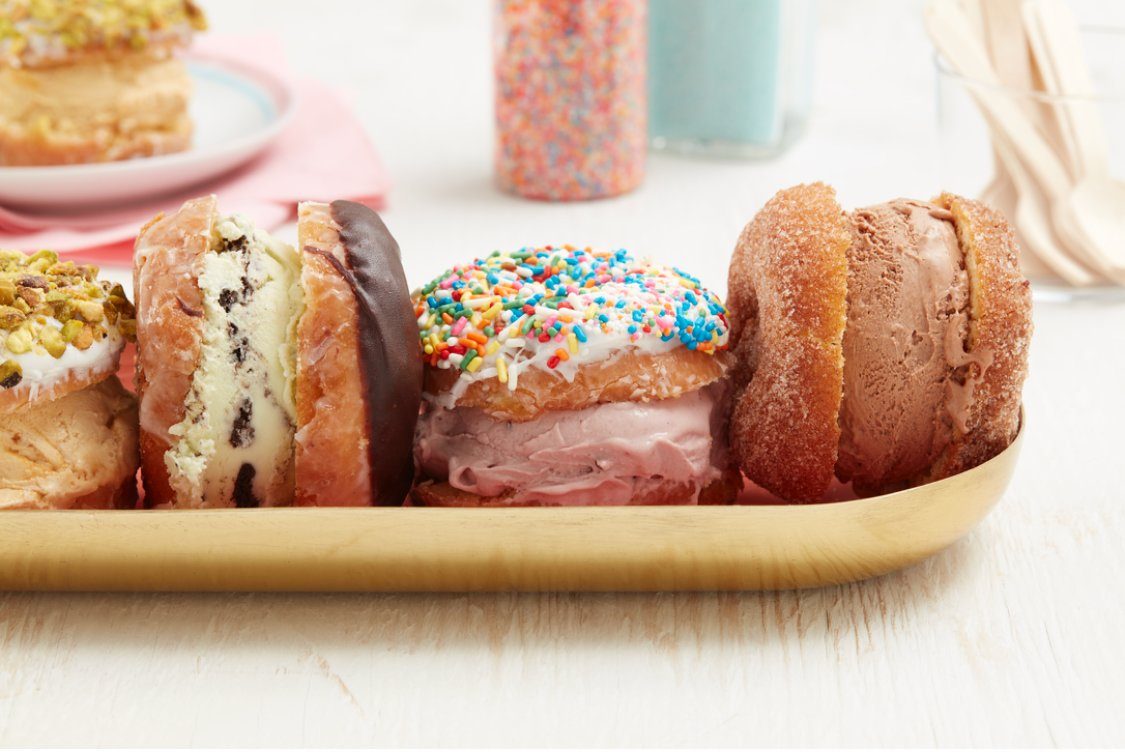 Ice cream sandwiches rest on a wooden tray, each made with donuts and varied ice cream flavors, with colorful sprinkles and toppings in a kitchen setting.