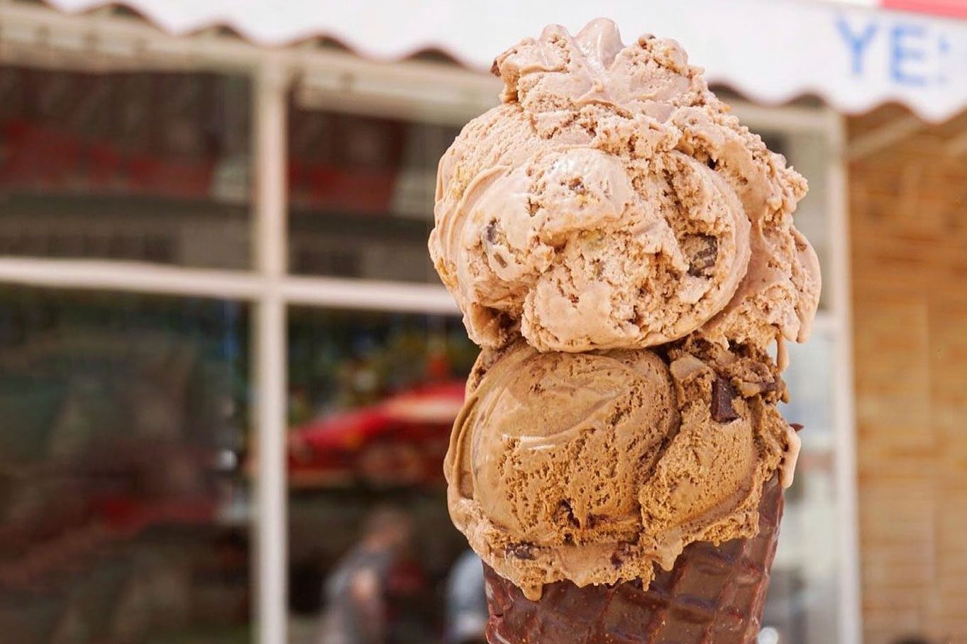 Scoops of chocolate ice cream piled on a waffle cone in front of a storefront with awning and glass windows in the background.