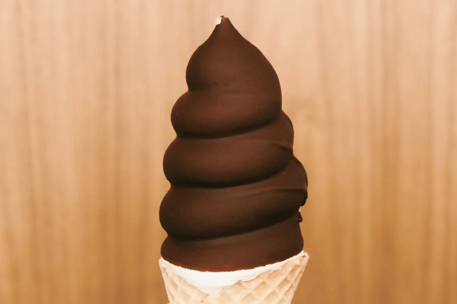 A chocolate-dipped soft serve ice cream cone sits against a light wood-textured background.