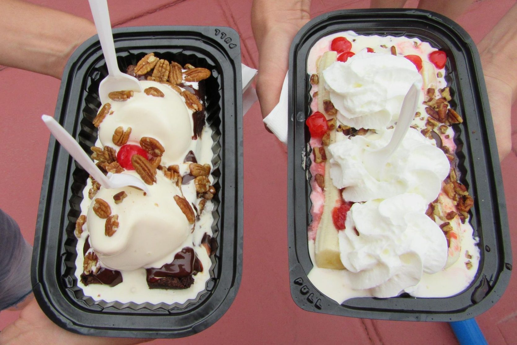 Two ice cream sundaes held in trays. Topped with pecans, cherries, and whipped cream. Background shows a red outdoor surface.