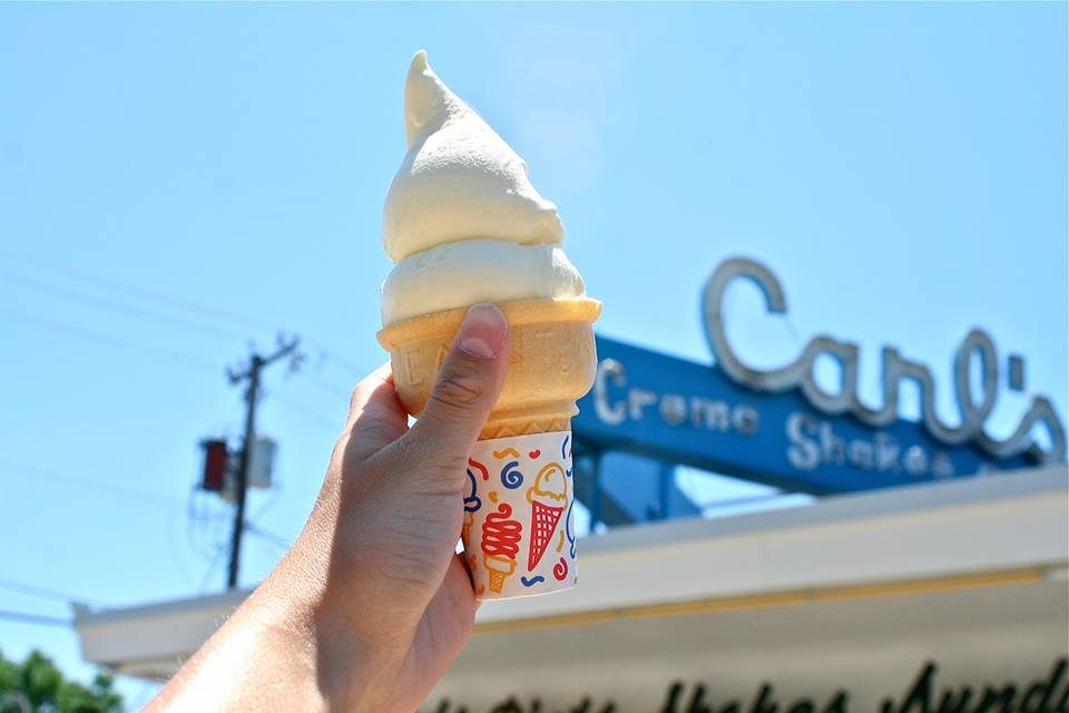 Hand holds vanilla ice cream cone against a clear sky, near a sign reading â€œCarl's.â€