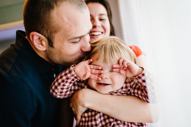 Child smiling, covering eyes, while being hugged. Adult kisses child's head, another smiles in the background. Bright indoor setting.
