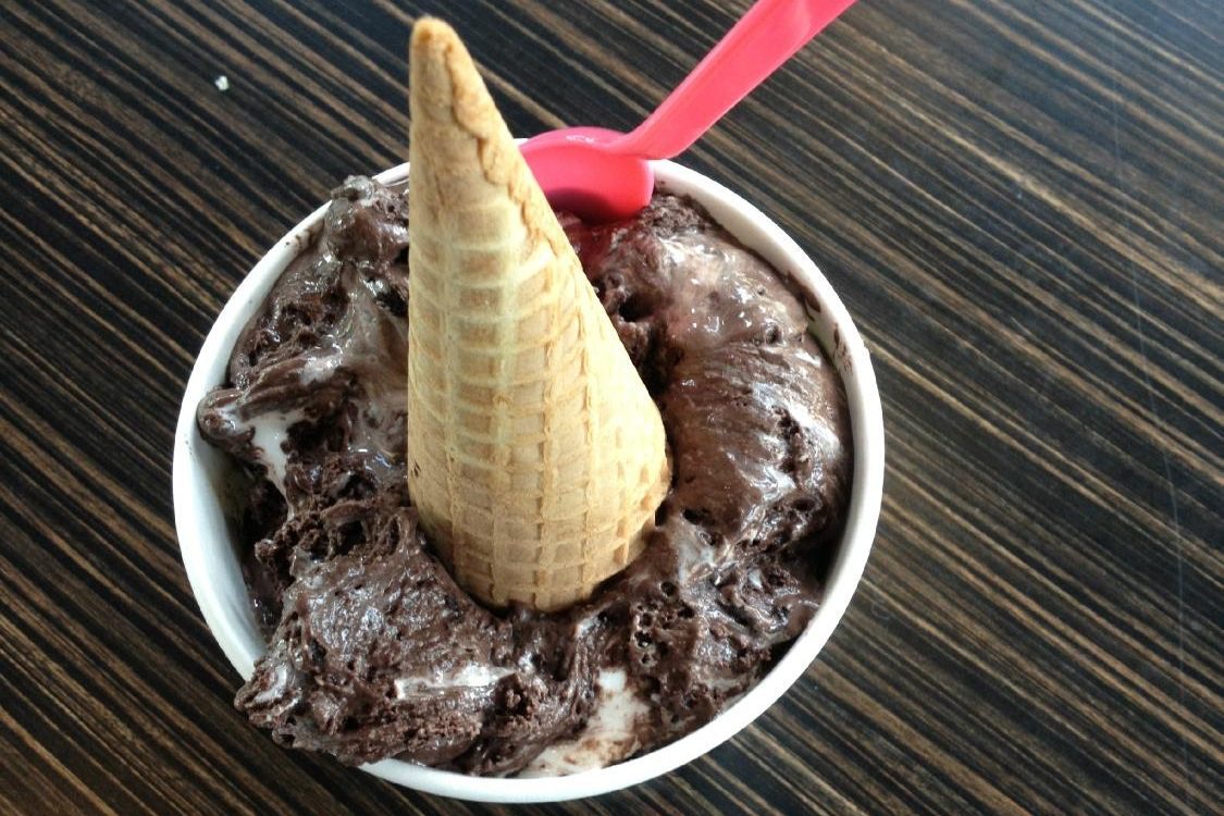 A waffle cone sits inverted in chocolate ice cream, served in a cup with a red spoon on a dark, striped table surface.