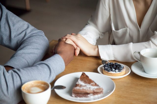 Hands clasped together across a wooden table with coffee and desserts, including a slice of cake and a blueberry tart.