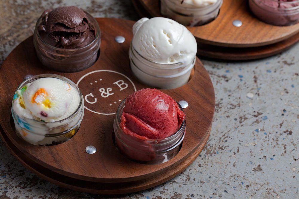 Small jars hold assorted ice cream flavors on a wooden tray with "C & B" engraved, placed on a textured gray surface.