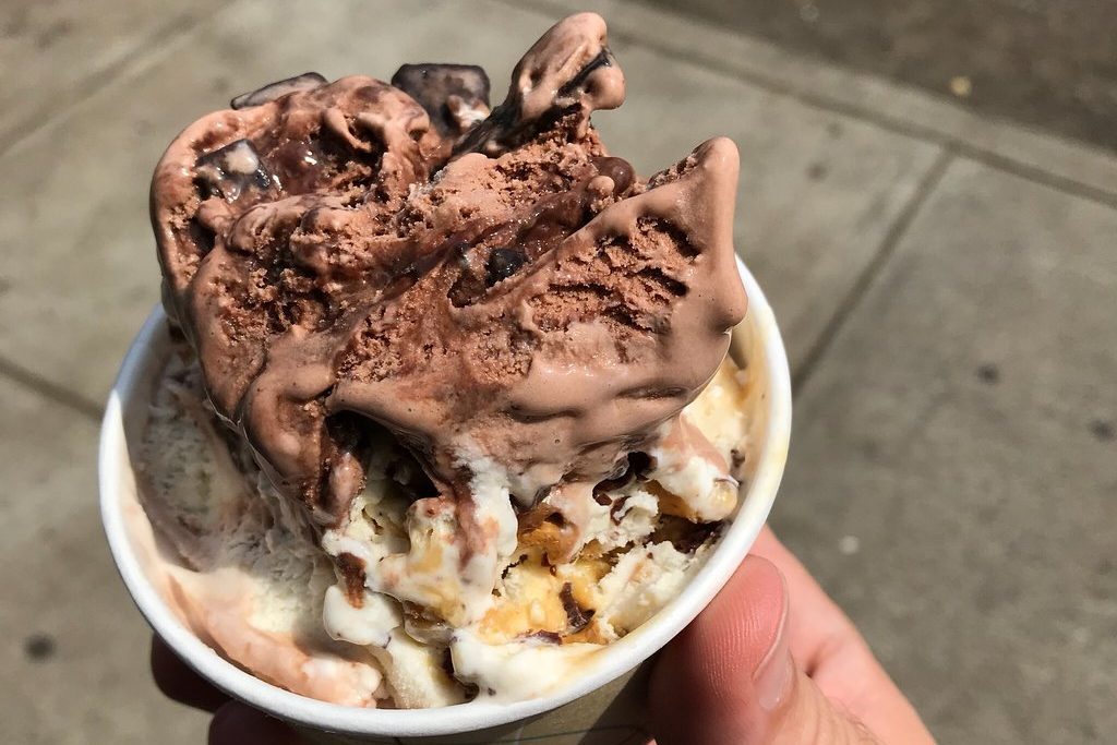 Chocolate and vanilla ice cream swirls, melting slightly, held in a paper cup by a hand on a sunlit sidewalk.