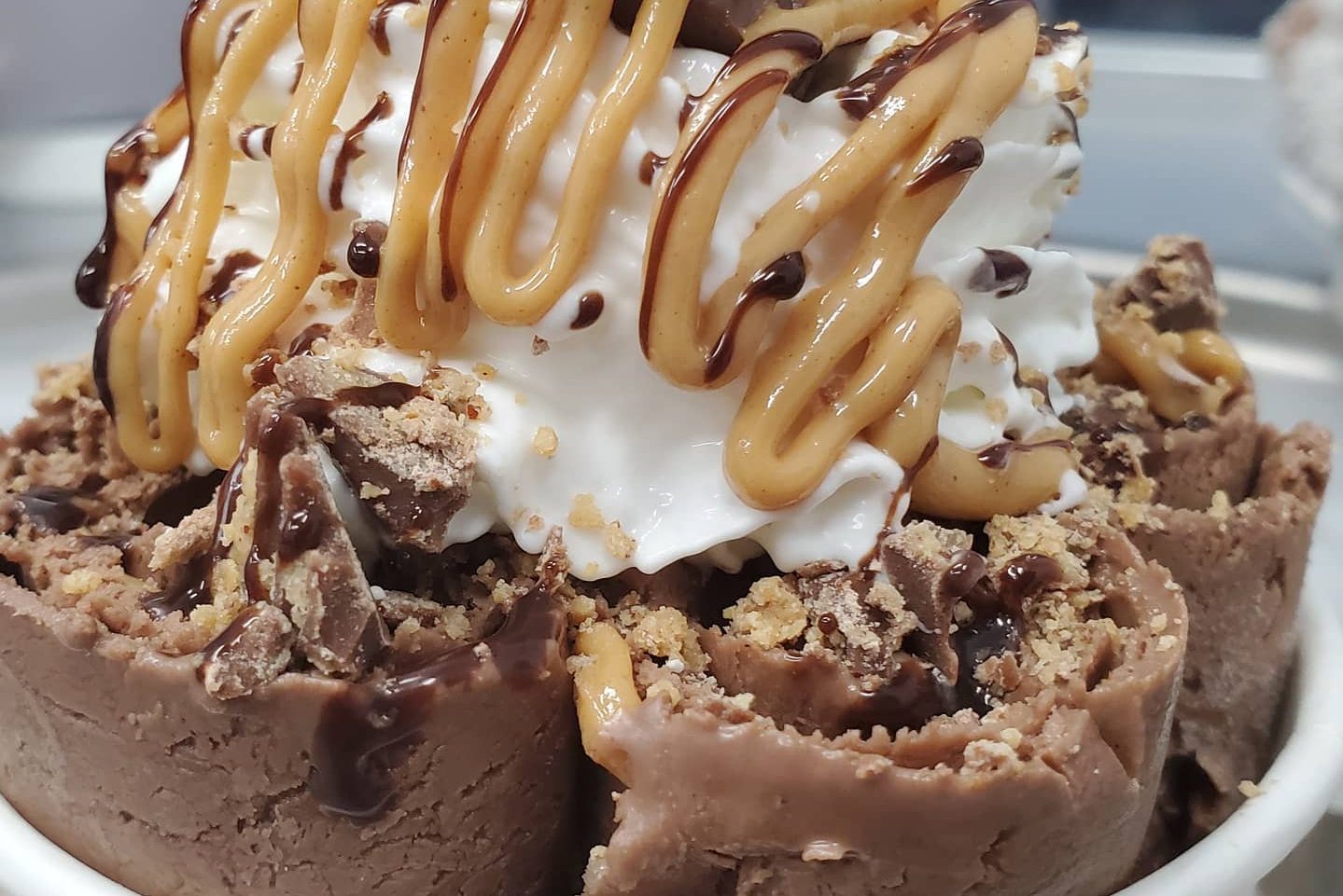 Chocolate rolled ice cream topped with whipped cream, peanut butter drizzle, and chocolate sauce, set in a white ceramic bowl.