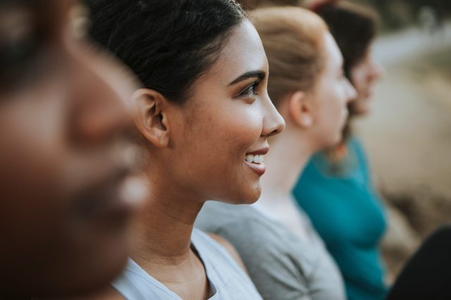 Four people seated in profile, smiling, in an outdoor setting. The focus is on the woman in the foreground.