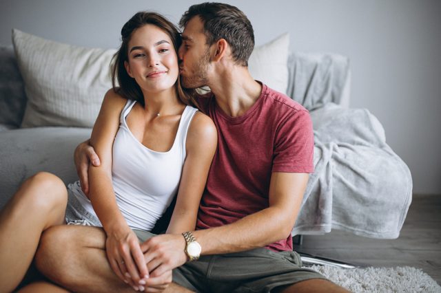 A couple sits on the floor, with the man kissing the womanâ€™s cheek, in a cozy room near a couch.