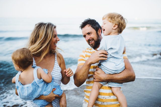 Family smiles, holds children, enjoys beach; waves gently crash in the background.