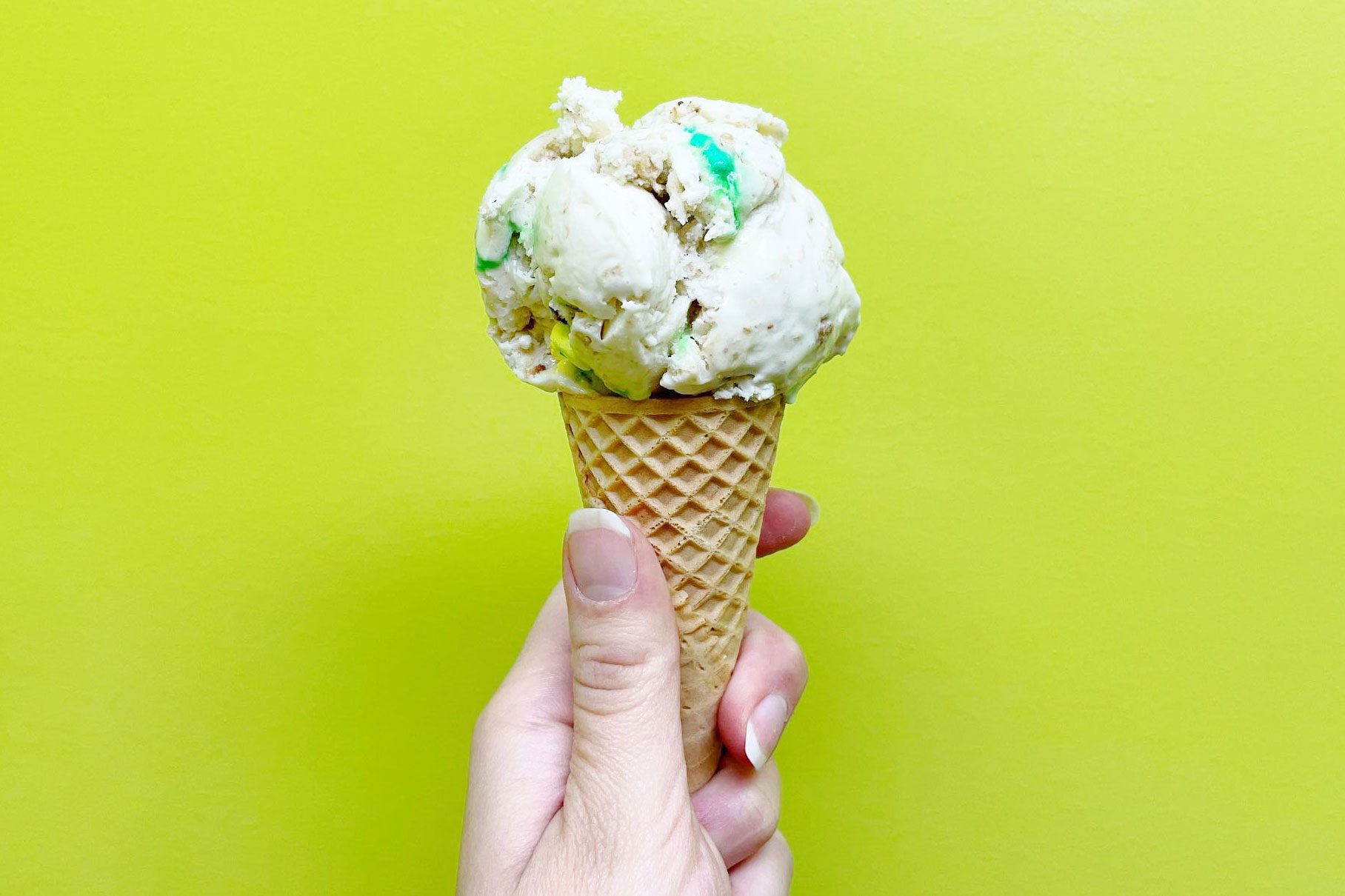 A hand holds an ice cream cone with multicolored scoops against a bright green background.