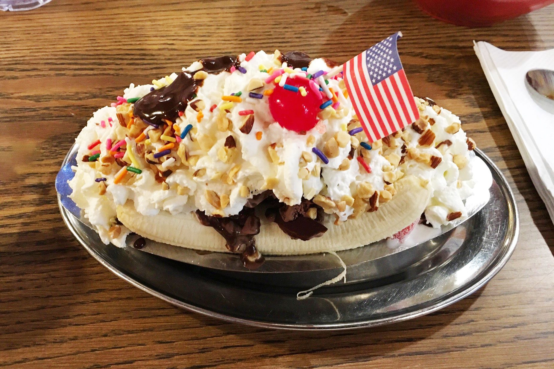 A banana split topped with whipped cream, cherries, nuts, and an American flag, sits on a wooden table, placed on a metal dish.