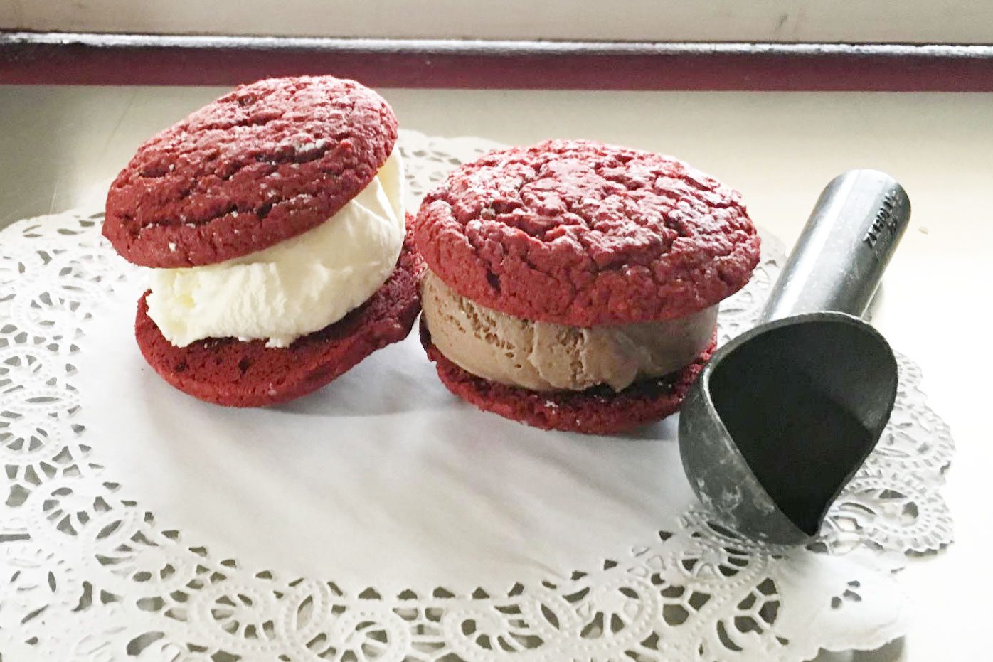 Ice cream sandwiches with red velvet cookies sit on a lace doily, accompanied by a metal ice cream scoop.