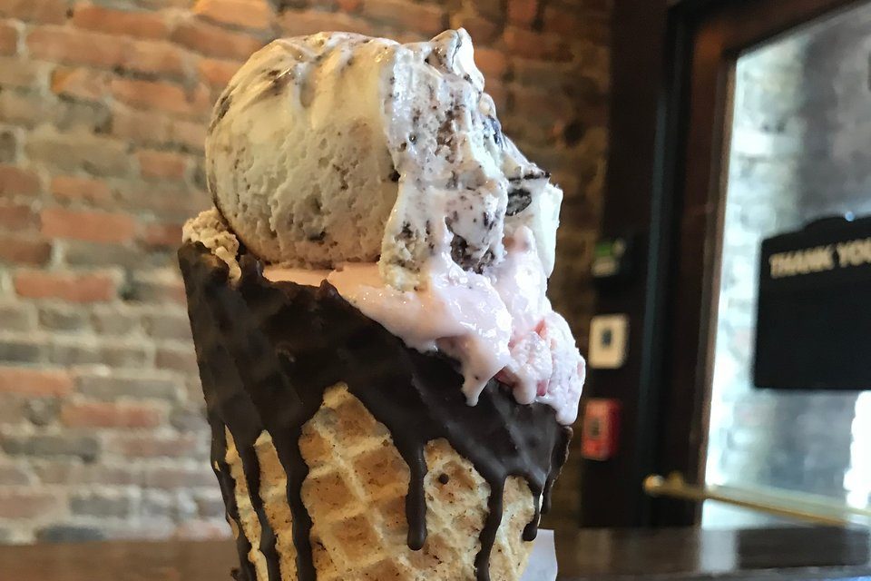 Ice cream drips from a waffle cone topped with chocolate syrup in a cozy cafÃ© with exposed brick walls.