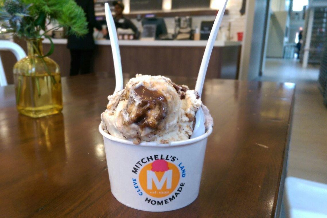 Ice cream sits in a cup on a table, featuring the logo "Mitchell's Homemade Ice Cream," with a cafe counter in the background.